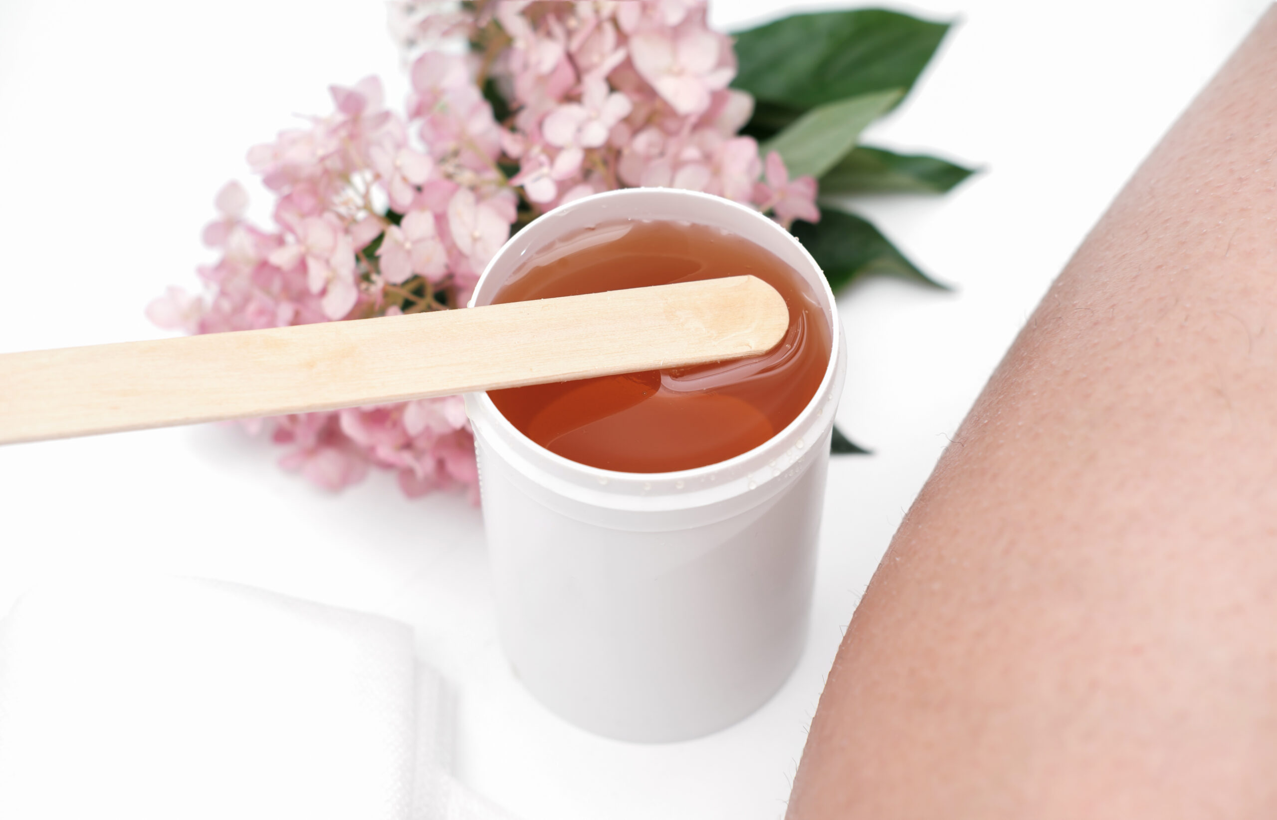 sugaring paste and spatula next to female leg. flowers on a background. depilation at home, skin and body care. hair removing. Why Waxing is Better for Your Skin